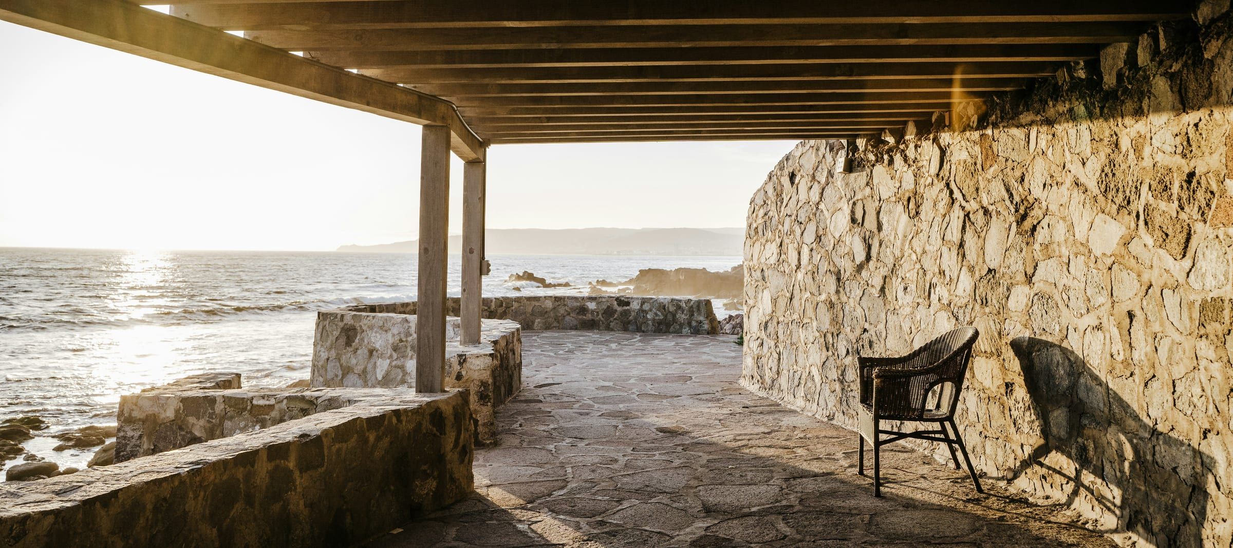 Terrasse couverte d'une pergola de bois, mur de pierre et fauteuil, face à la mer en contre-jour